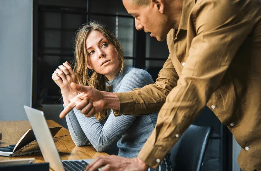 Homem e mulher no escritório, com o homem apontando para o laptop enquanto a mulher senta pensativa, sugerindo orientação ou suporte profissional em uma conversa de trabalho