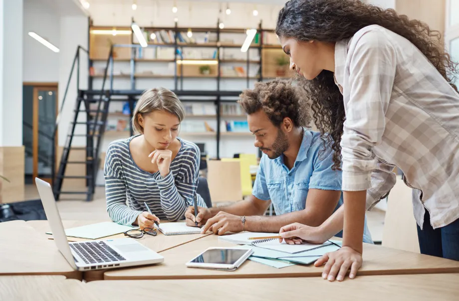 Equipe em ambiente de escritório trabalhando em um projeto: uma pessoa desenha e aponta em papéis, enquanto outras utilizam notebook e celular, com atenção em tarefas de trabalho.