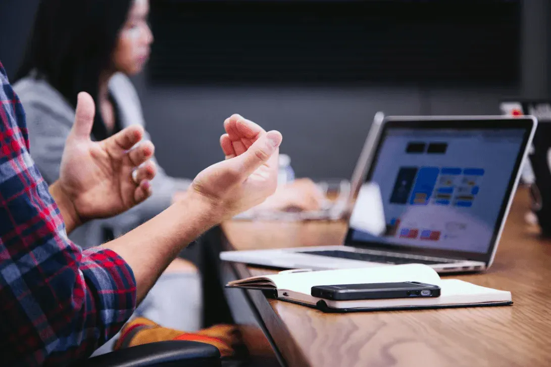 Pessoa em reunião aponta para um laptop com gráficos e indicadores na tela, enquanto discute ideias com outras pessoas ao fundo em ambiente de trabalho