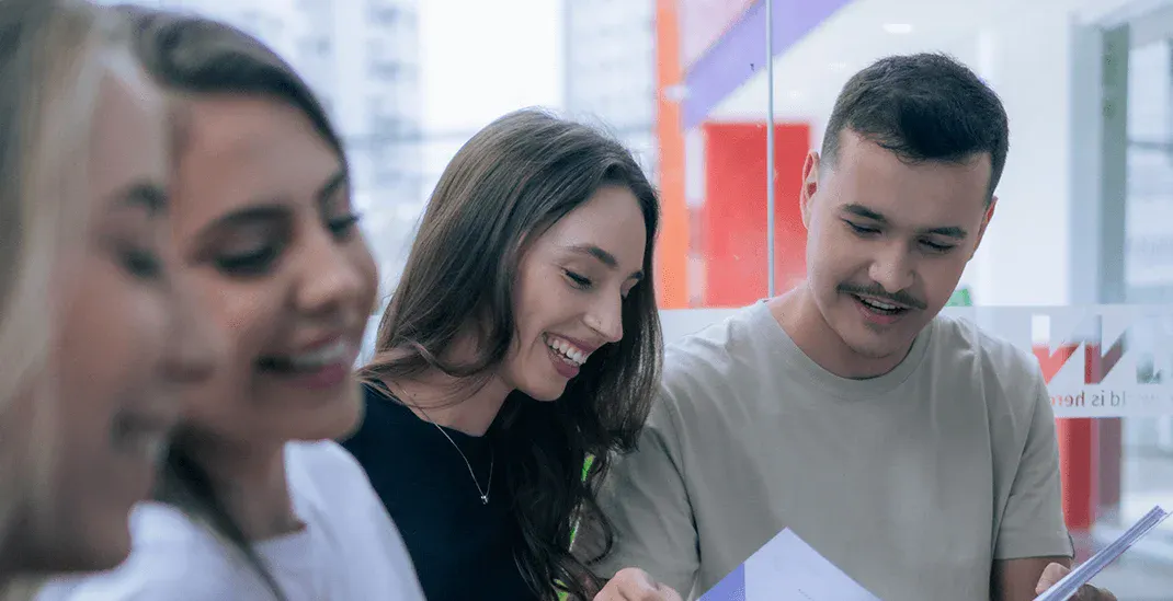 Três pessoas em um ambiente de escritório analisando documentos e compartilhando ideias, com sorrisos durante uma reunião ou treinamento em equipe.
