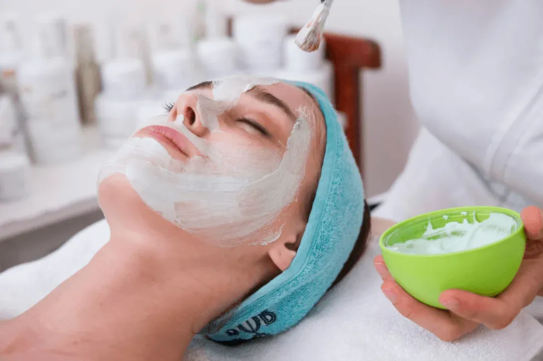 Spa facial com máscara cremosa branca aplicada no rosto de uma mulher, com touca descartável azul, pincel em uso e tigela verde com produto cosmético ao lado em ambiente de tratamento