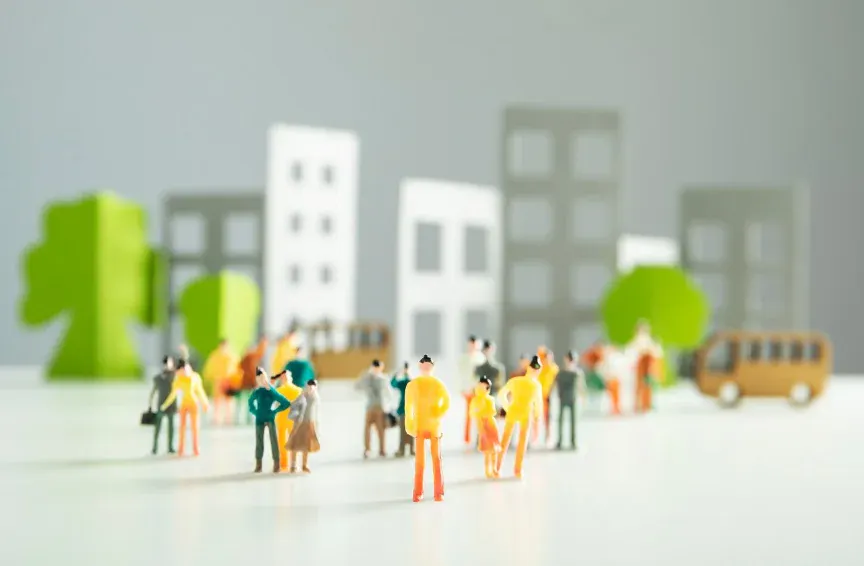 Cena em miniatura com bonecos em escala, prédios coloridos ao fundo e pessoas caminhando em uma cidade, com aparência de modelo urbano.