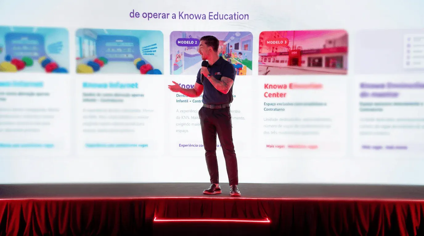 Palestrante em pé em um palco durante apresentação em evento educacional, ao fundo painéis com informações e a marca Knowa Education exibida em cartões de modelo.