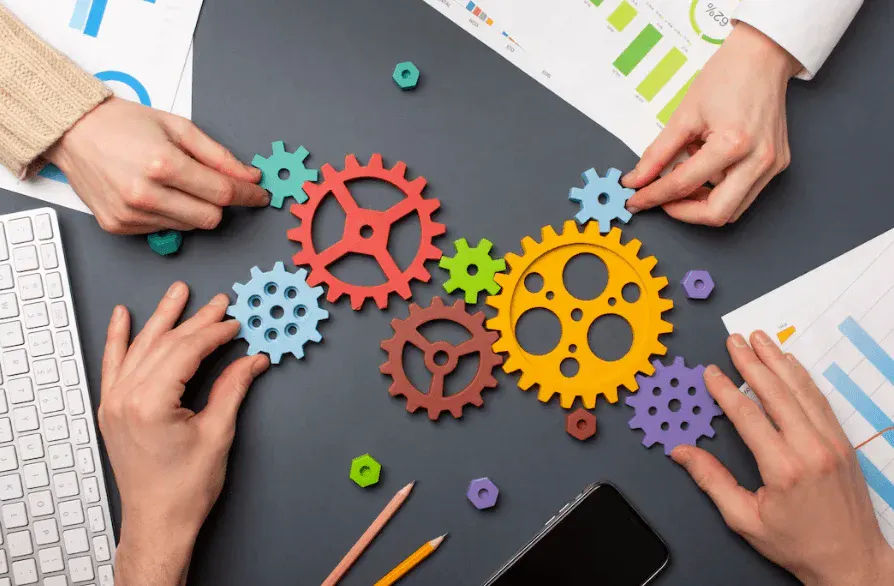 Mãos colaborando sobre uma mesa com engrenagens coloridas e ícones de gestão de projetos, enquanto ferramentas e documentos aparecem ao redor, representando trabalho em equipe e planejamento estratégico.