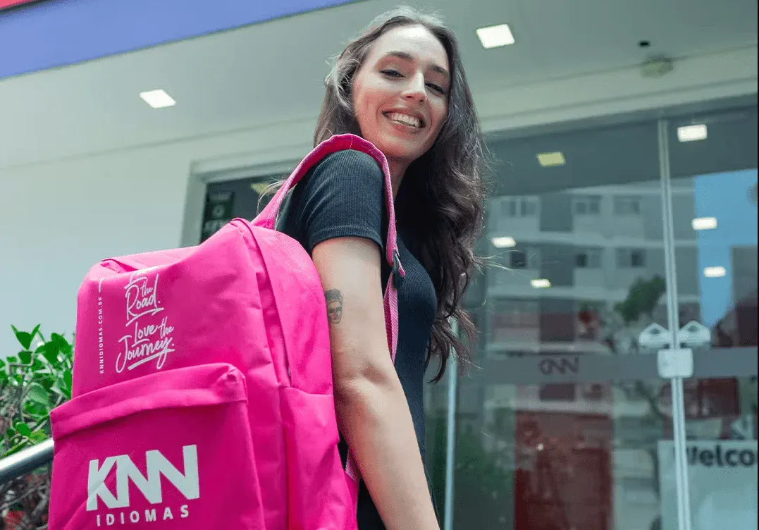 Mulher sorridente carregando uma grande sacola rosa com o logotipo “KNN Idiomas” em frente a uma fachada de escola ou escritório. A sacola está aberta no braço e o fundo mostra janelas com reflexos.