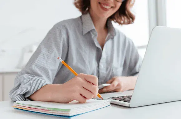 Mulher sentada em uma mesa, usando laptop e fazendo anotações com lápis em um caderno, em ambiente de trabalho ou estudo.