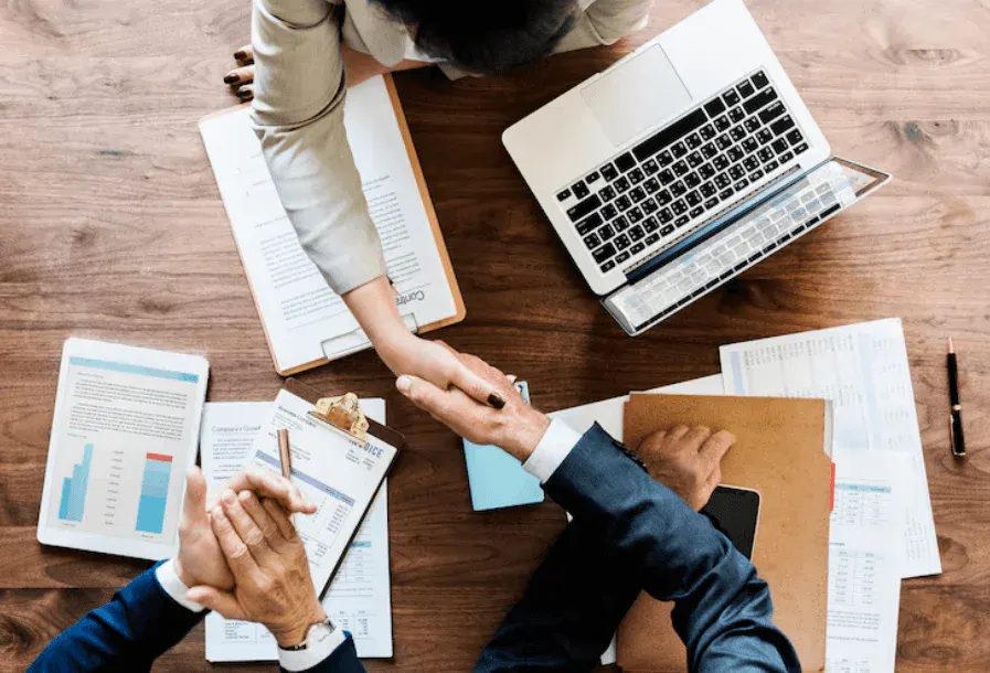 Reunião de trabalho em mesa de madeira: pessoas analisam relatórios e documentos, enquanto um notebook está aberto ao lado de planilhas, sugerindo planejamento e gestão de projetos.
