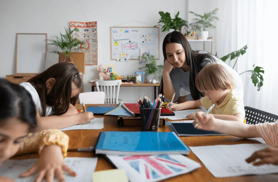 Professora e crianças estudando e desenhando em sala de aula, com materiais escolares e placas ao fundo, em atividade educativa em grupo.