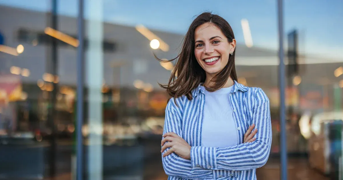 Mulher sorrindo com braços cruzados em frente a uma vitrine de loja, usando camisa listrada azul e branca, com fundo desfocado de ambiente comercial.