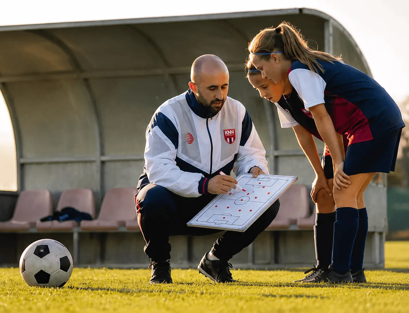 Treinador e jogadora analisam um quadro tático com marcações em um campo de futebol ao ar livre, em treino na grama