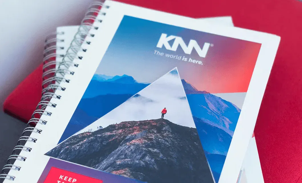 Cadernos abertos com capa do KMV, com imagens de montanhas e paisagens, sobre fundo vermelho; destaque para design gráfico com formas e logotipo KNN.