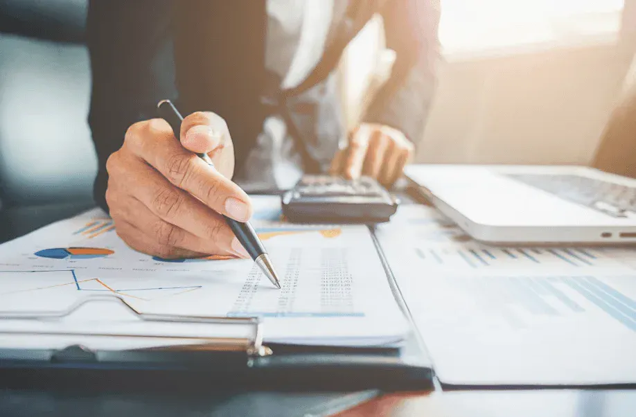 Pessoa escrevendo e fazendo anotações em documentos com gráficos de planejamento e dados financeiros sobre uma mesa, enquanto um laptop e calculadora ficam ao fundo, em ambiente corporativo