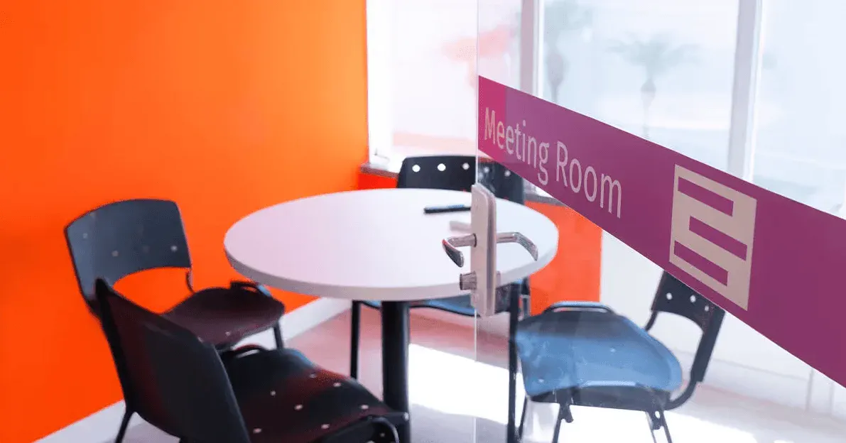Sala de reunião moderna com mesa redonda branca e cadeiras escuras, placa de sinalização “Meeting Room” na divisória e ambiente corporativo com cores vibrantes.