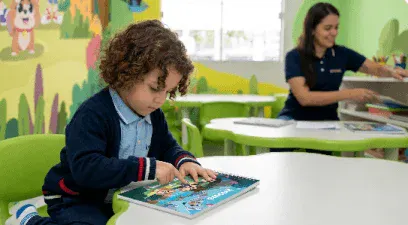Criança em sala de aula escolar usando um livro ilustrado sobre uma mesa verde, enquanto uma professora trabalha ao fundo.