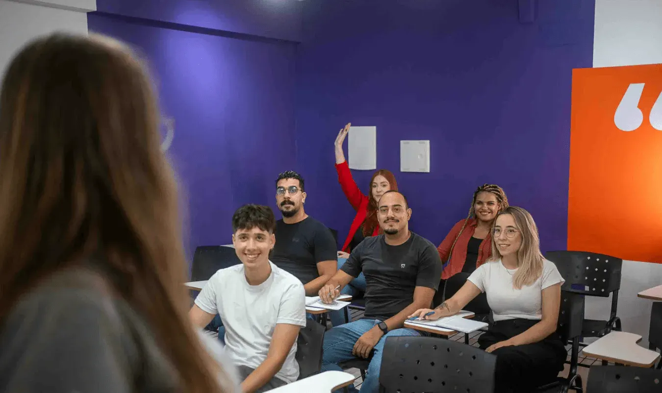 Equipe de pessoas em sala de treinamento com cadeiras e mesas, discutindo com atenção, enquanto uma pessoa ao fundo levanta o braço e todos parecem participar de uma atividade em grupo.