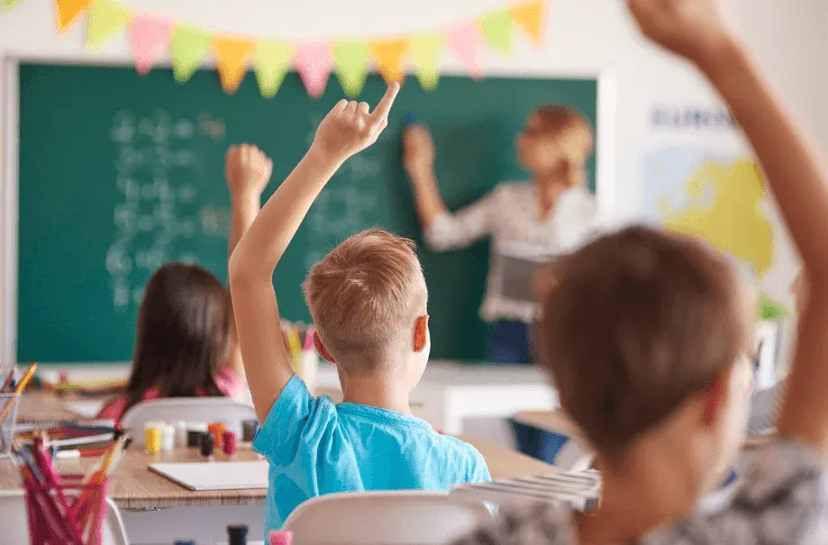 Criança em sala de aula aponta para a lousa verde, enquanto o professor fala ao fundo e outros alunos assistem. Ambiente escolar com decoração colorida.