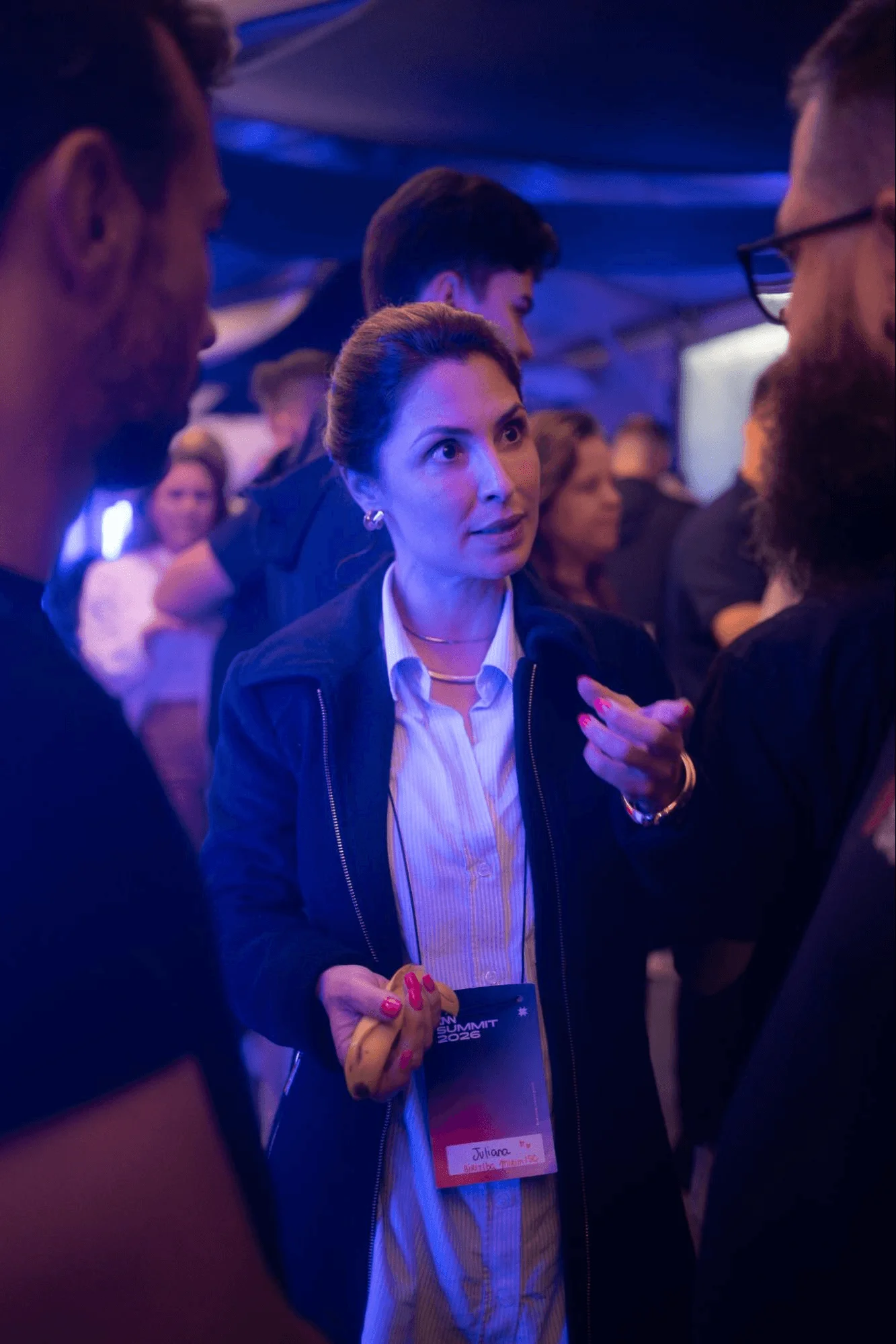 Mulher em evento corporativo em ambiente interno, usando credencial com a inscrição “SUMMIT 2026” e “Jathane”, iluminada por luz azul e conversando com pessoas ao redor.