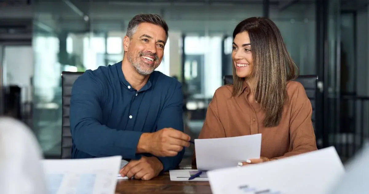 Dois profissionais em um escritório moderno conversam e sorriem durante uma reunião, com papéis e documentos sobre a mesa, sugerindo negociação ou apresentação de propostas.