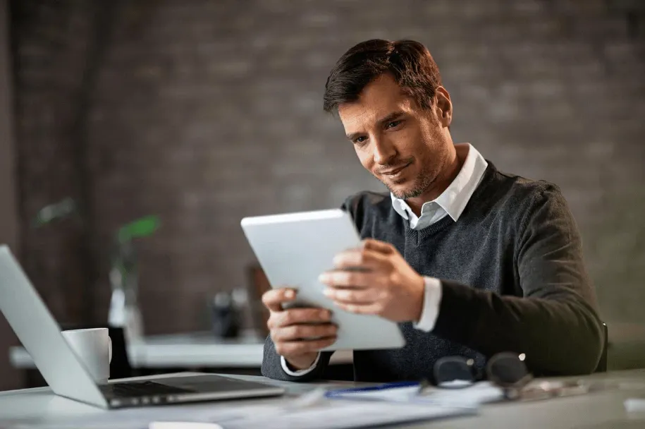Homem trabalhando em escritório moderno, usando um tablet e um notebook sobre a mesa, em ambiente corporativo com fundo desfocado