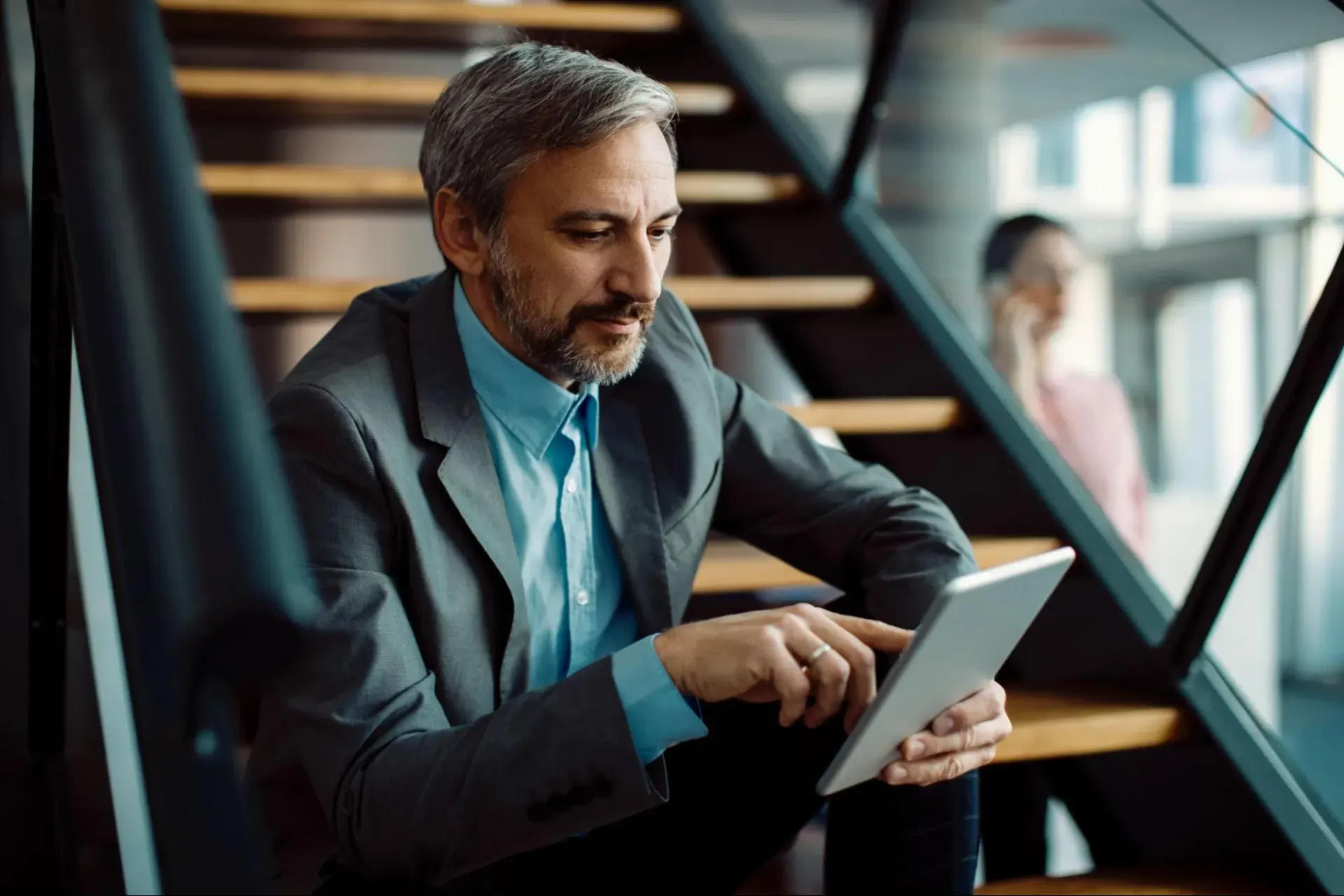 Homem de meia-idade com barba e cabelo grisalho, em ambiente corporativo, usando tablet e consultando informações enquanto trabalha em um escritório com escadas ao fundo.