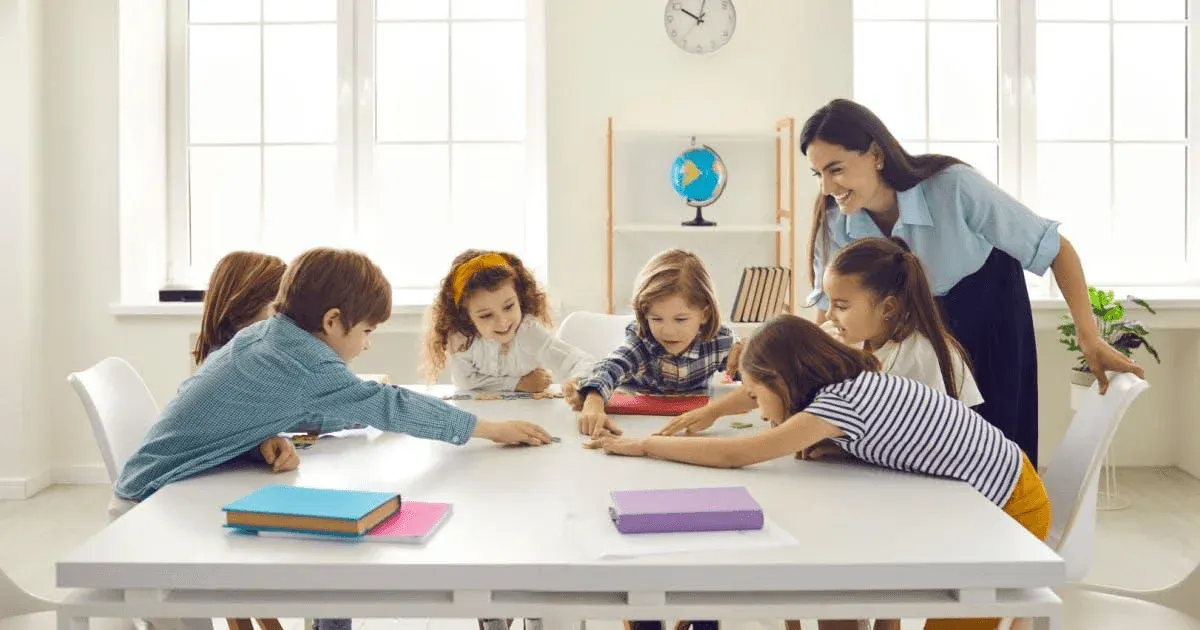 Sala de aula com professora sorridente orientando crianças sentadas à mesa, enquanto elas usam livros e fazem atividades educativas perto de um globo e relógio na parede.