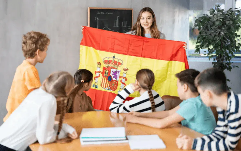Professora em sala de aula apresenta a bandeira da Espanha para crianças durante atividade educativa, com quadro ao fundo e materiais sobre a mesa