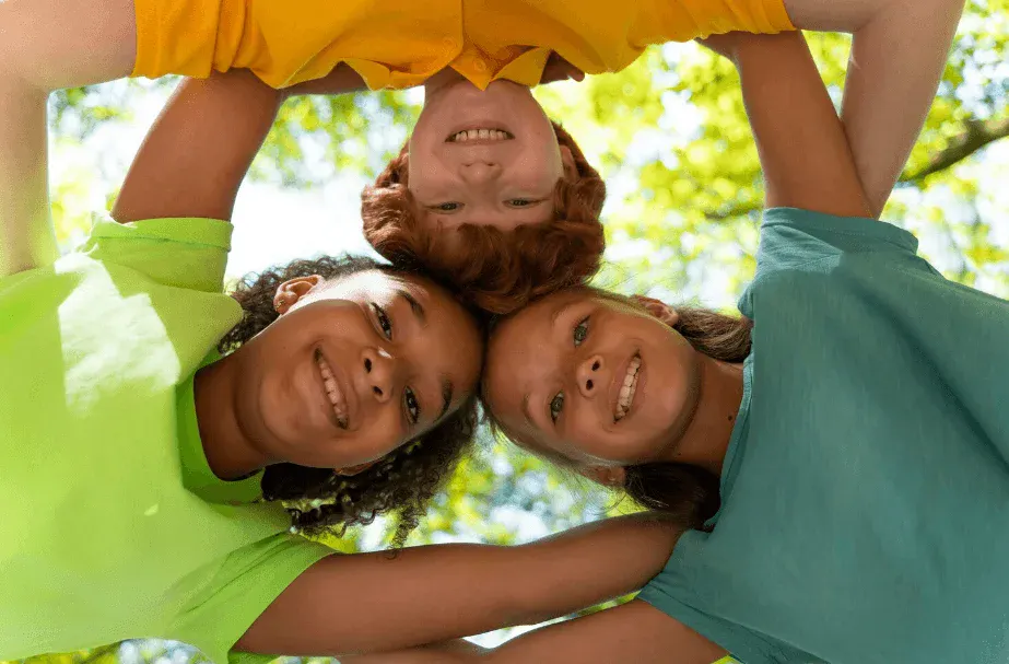 Foto de três crianças sorrindo e abraçadas, fazendo uma brincadeira ao ar livre, com fundo desfocado de árvores e luz natural em dia ensolarado.