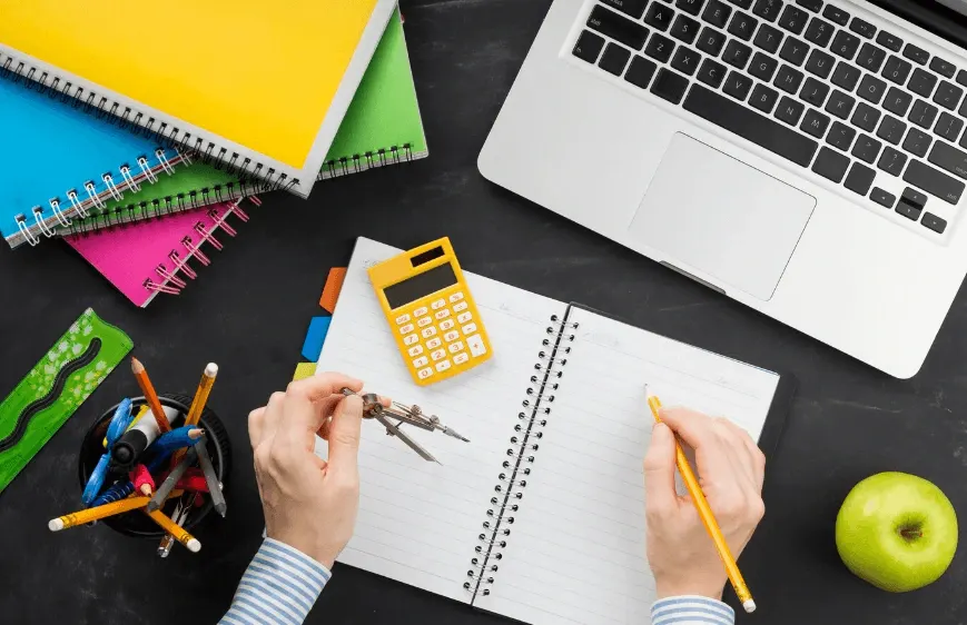 Pessoas estudando e planejando tarefas com caderno aberto, lápis e calculadora em uma mesa com apple, lápis de cor e material escolar, ao lado de um notebook