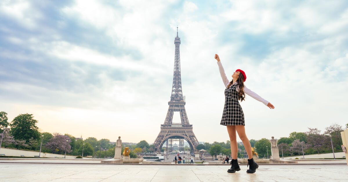 Uma mulher com vestido e chapéu está em pé em frente à Torre Eiffel.