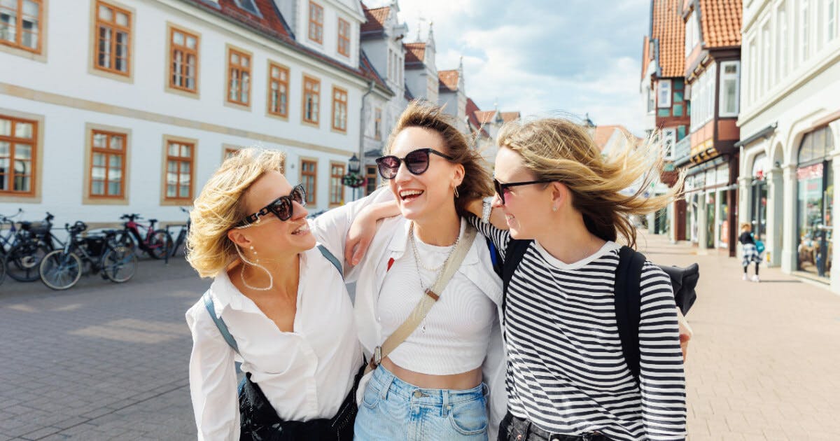 Três jovens mulheres caminhando pela rua, conversando e sorrindo, em um ambiente urbano.