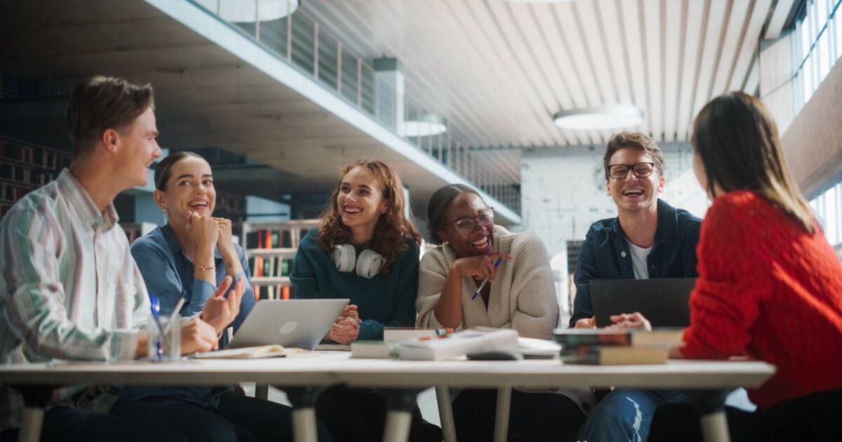 Grupo de jovens sentados à mesa em uma biblioteca, envolvidos em discussões e estudos.