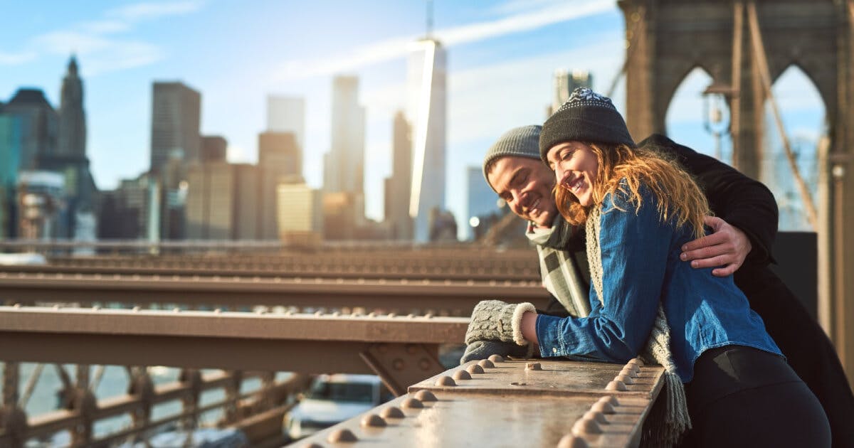 Casal caminhando sobre a Ponte do Brooklyn, em Nova York, com a cidade ao fundo e um céu claro.