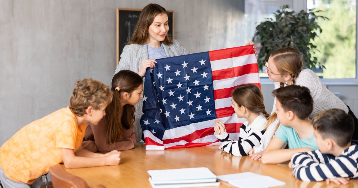 Professor segurando uma bandeira americana em frente a um grupo de crianças, simbolizando patriotismo e educação.