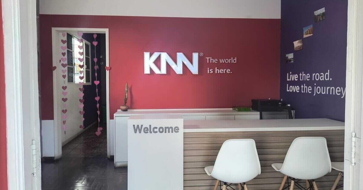 Entrada da KNN Idiomas Esmeraldas no estado de Minas Gerais. Na foto tem a parede com a logo da KNN, uma bancada escrito "Welcome" e duas cadeiras brancas.