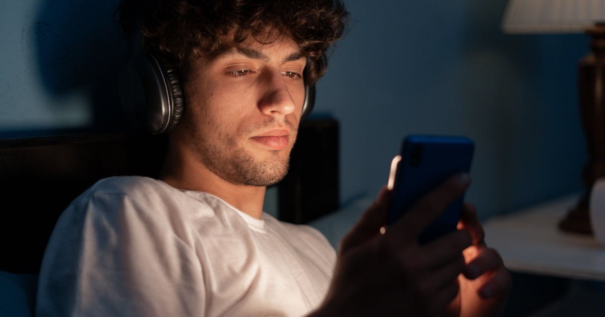 Um jovem de fone de ouvido olhando para um celular deitado na cama.