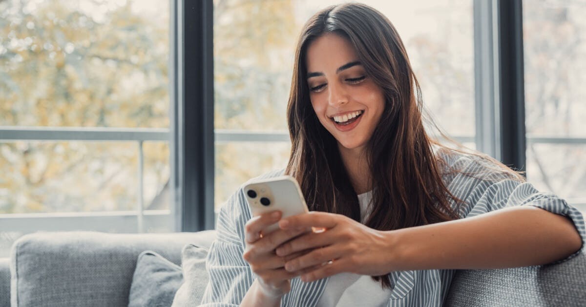 Jovem de cabelo preto e comprido, sorrindo e olhando para a tela de seu celular.