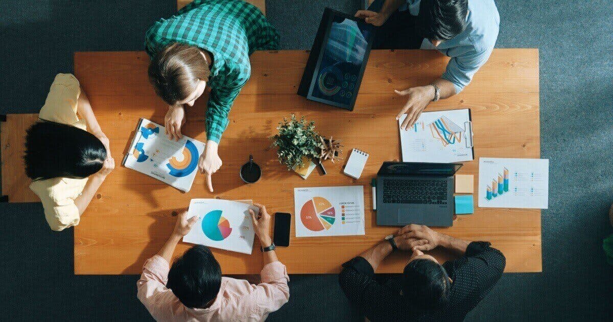 Imagem aérea de 5 pessoas em uma sala de reunião e com folhas mostrando infográficos.