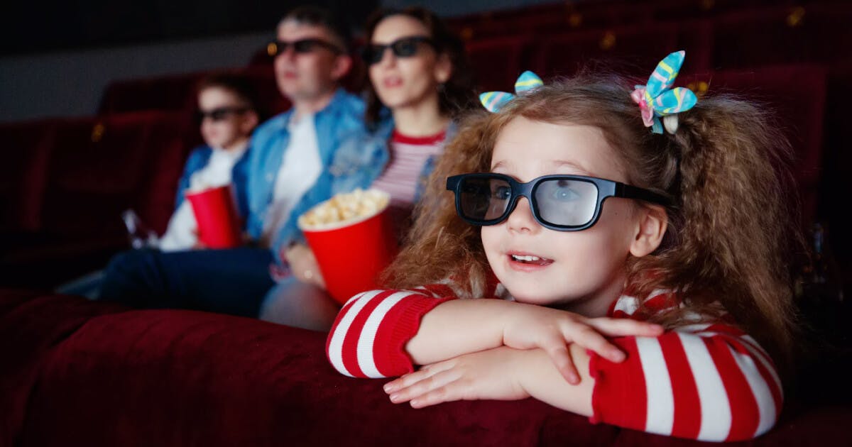 Menina pequena usando óculos 3D, sentada em um teatro, assistindo a um filme com expressão de encantamento.