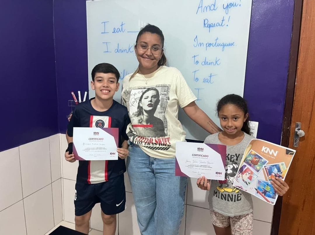 Professora da KNN sorrindo ao lado de dois alunos na KNN Idiomas Manhumirim, em MG.