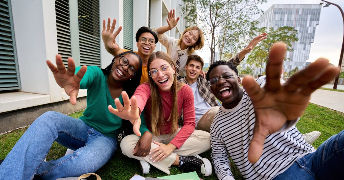 Grupo de jovens sentados na grama, com as mãos levantadas, expressando alegria e união em um momento de descontração.