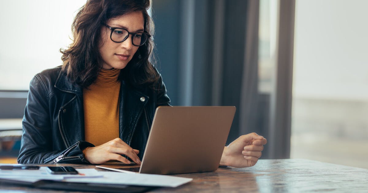 Uma mulher de óculos sentada à mesa com um laptop, concentrada em seu trabalho.