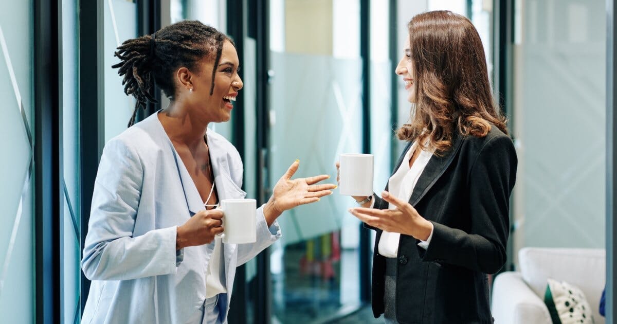 Duas mulheres, uma com blazer azul marinho e outra com blazer preto, conversando e cada uma com uma xícara de café.