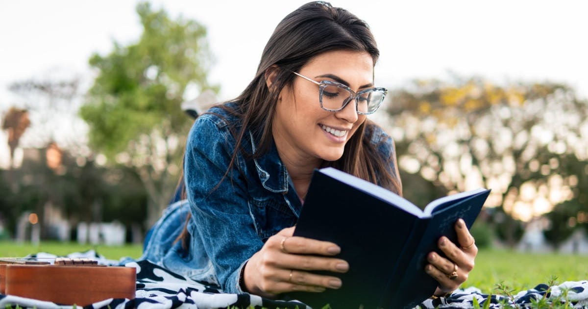 Mulher de óculos deitada na grama, sorrindo e com um livro na mão.