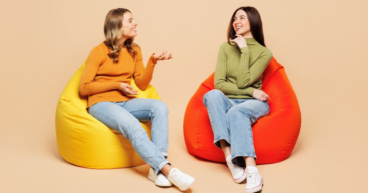 Duas mulheres sorrindo olhando uma para a outra sentadas em puffs.