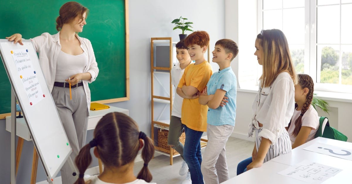 Professora apontando para um quadro na frente de alunos crianças.