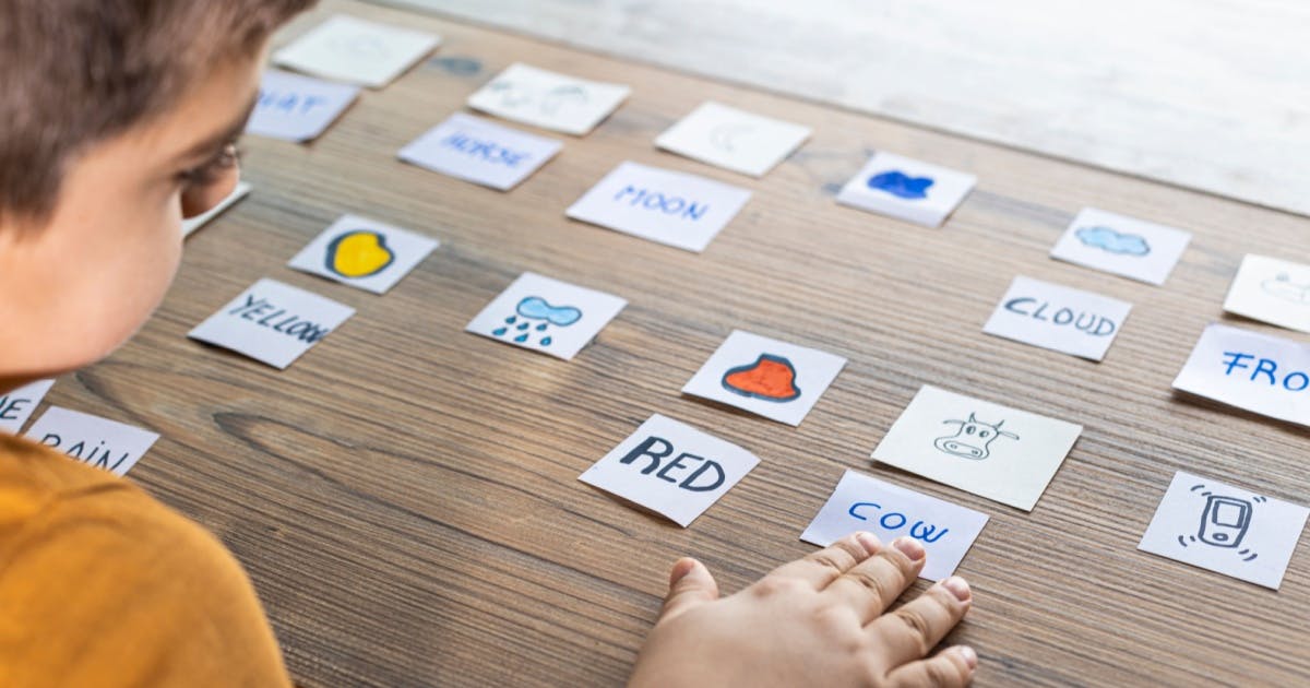 Criança colando papéis com nome de cores e objetos em um quadro.