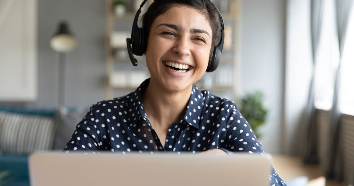 Mulher dando risada na frente de um notebook e com um headset no ouvido.