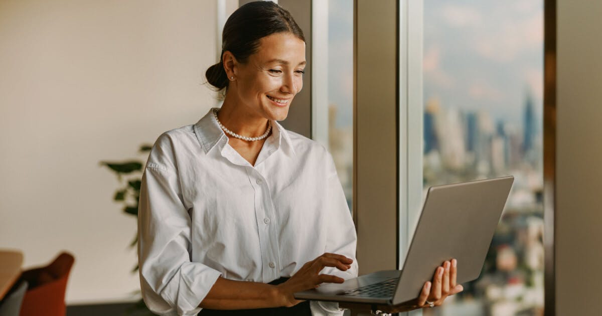 Uma mulher com camisa branca segura um laptop, olhando para a tela com expressão concentrada.