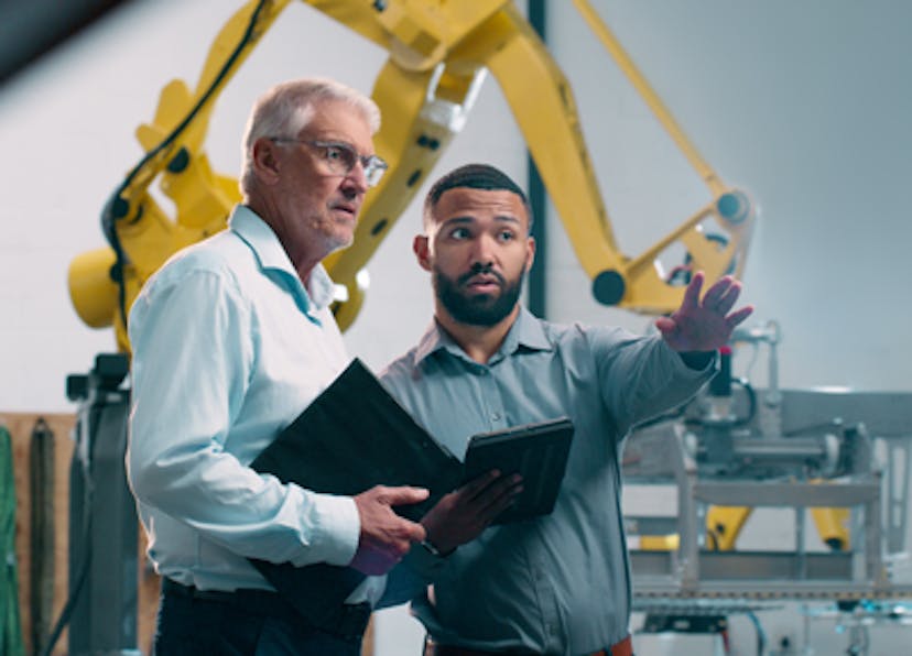 två-personer-inom-iot-ai-automation-pratar-med-varandra-i-industrimiljo-robotar-bakgrunden_mdr