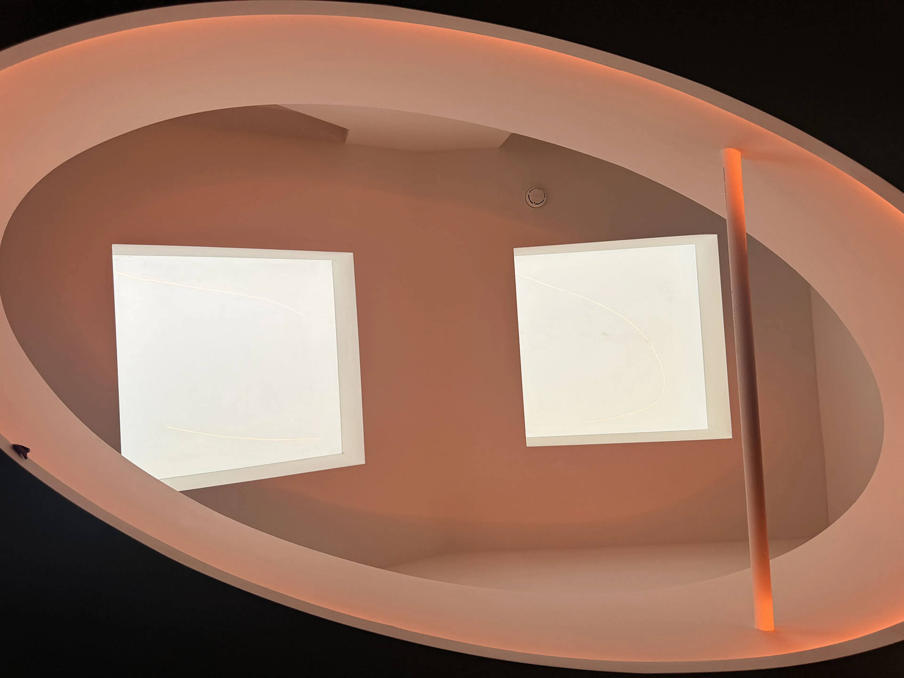 Oval architectural ceiling recess backlit with warm orange LED strip lighting and square skylight elements